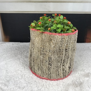 May include: A cylindrical woven fabric planter featuring a textured beige and black pattern with a bright pink stitched trim along the top and bottom edges, containing a cluster of green artificial foliage with small orange flower buds.