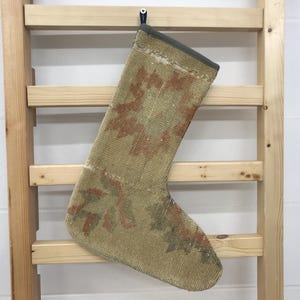 May include: A beige Christmas stocking with a floral pattern in shades of red and green. The stocking is made of a woven material and has a dark gray cuff. It hangs on a wooden ladder against a white wall.