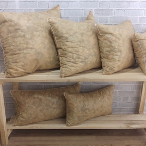 May include: Six beige throw pillows with a subtle pattern are displayed on a wooden shelf unit. The pillows are arranged in two rows, with three pillows in each row.