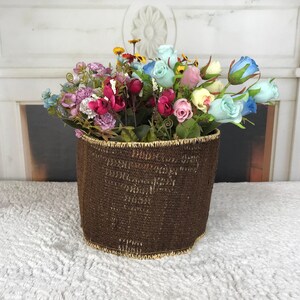 May include: A dark brown woven basket with a light tan rim filled with an assortment of artificial flowers in shades of light blue, pink, yellow, and purple, featuring green leaves and stems arranged within the cylindrical container.