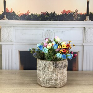 May include: A bouquet of colourful artificial flowers in a rustic, woven, cream-coloured planter. The planter is sitting on a wooden table in front of a white marble fireplace.