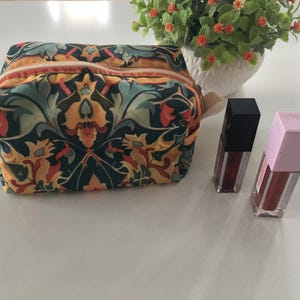 Puede incluir: Un neceser con estampado floral con cremallera beige, junto a dos tubos de brillo labial. La bolsa presenta un diseño en tonos turquesa, naranja y amarillo. Un pequeño jarrón blanco con flores verdes y naranjas está en el fondo.