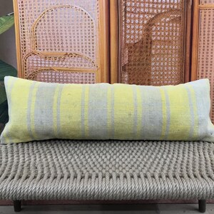 Puede incluir: Un cojín lumbar rectangular con rayas verticales amarillas y grises. El cojín está colocado sobre un banco tejido. El fondo presenta una pantalla de madera y una gran planta verde.