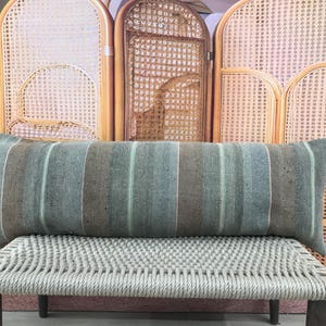 Peut inclure: Un long coussin rectangulaire à rayures verticales dans les tons de bleu, marron et blanc. Le coussin repose sur un banc tressé. L'arrière-plan présente un paravent décoratif en bois et une plante en pot.