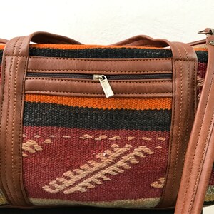 Op de afbeelding: Een bruine leren en geweven tapijt duffelbag met een ritsvak en een lange band. De tas heeft een kleurrijk geometrisch patroon in tinten rood, oranje, zwart en roze.