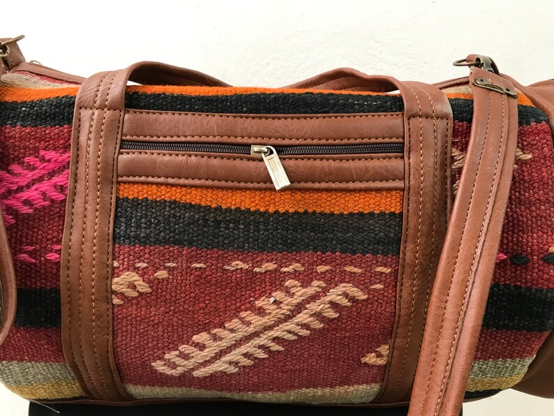 Op de afbeelding: Een bruine leren en geweven tapijt duffelbag met een ritsvak en een lange band. De tas heeft een kleurrijk geometrisch patroon in tinten rood, oranje, zwart en roze.
