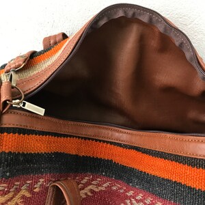 Op de afbeelding: Een bruine leren reistas met een ritssluiting en een kleurrijk geweven patroon. De tas is open en de binnenkant is gevoerd met bruin stof.