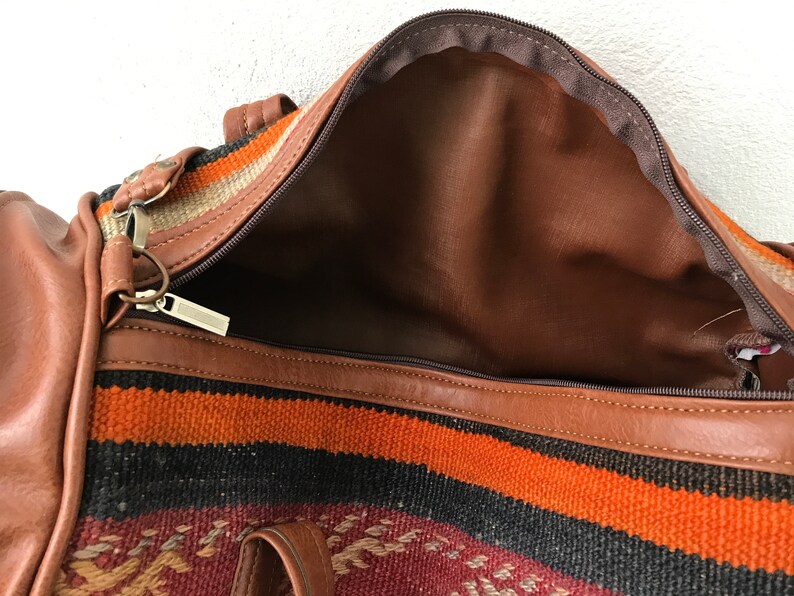 Op de afbeelding: Een bruine leren reistas met een ritssluiting en een kleurrijk geweven patroon. De tas is open en de binnenkant is gevoerd met bruin stof.