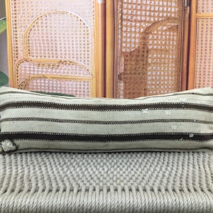 Pode incluir: Almofada retangular com listras horizontais marrons sobre um fundo bege claro. A almofada é colocada sobre uma superfície tecida. O design é simples e rústico, adequado para decoração de casa.