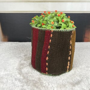 Puede incluir: Una maceta cilíndrica de tela con textura tejida y rayas verticales en tonos marrón oscuro, rojo intenso y amarillo mostaza. Los bordes están rematados con puntadas de festón en verde claro y contiene un grupo de pequeñas hojas verdes.