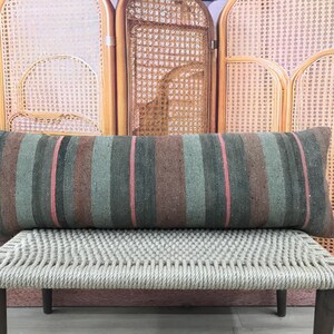 Puede incluir: Cojín rectangular con rayas verticales en tonos marrón, verde y rosa. El cojín está colocado sobre un banco tejido con patas de madera, con un biombo de mimbre y una planta en maceta de fondo.