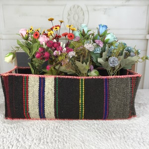 Può includere: Un cesto rettangolare intrecciato con un bordo rosa, pieno di una varietà di fiori artificiali colorati, tra cui rosa, rosso, giallo, blu e verde.