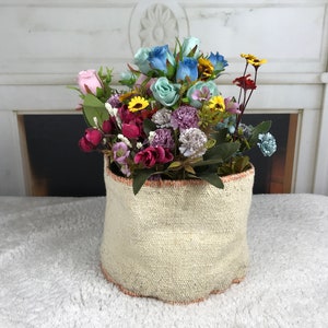 May include: A white woven basket with a pink, blue, and yellow floral arrangement. The basket has a brown trim around the top.