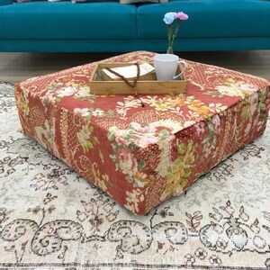 Pode incluir: Um pufe quadrado, vermelho e com padr&atilde;o floral, est&aacute; sobre um tapete de cor creme com design floral. O pufe est&aacute; na frente de um sof&aacute; azul turquesa.