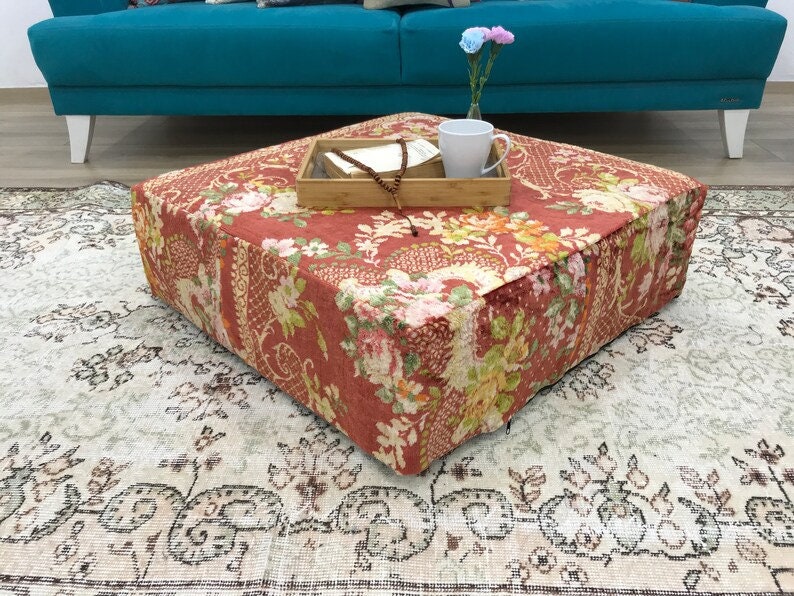Pode incluir: Um pufe quadrado, vermelho e com padr&atilde;o floral, est&aacute; sobre um tapete de cor creme com design floral. O pufe est&aacute; na frente de um sof&aacute; azul turquesa.
