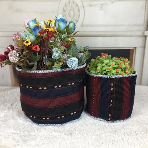 May include: Two woven baskets with blue, red and black stripes. The larger basket holds a bouquet of colourful artificial flowers, including blue, pink, yellow and red. The smaller basket holds a green plant with small orange flowers.