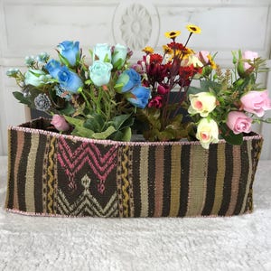 Puede incluir: Una cesta rectangular de tela con un patrón tejido de rayas marrones, bronceadas y rosas con motivos geométricos. La cesta está llena de un surtido de flores artificiales, incluyendo rosas azul claro, rosas rosadas y pequeñas margaritas amarillas.