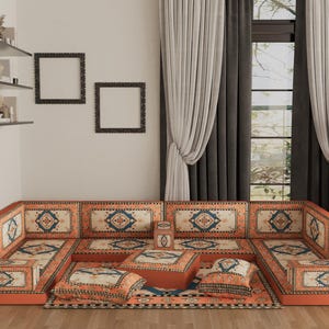 Könnte beinhalten: Ein orangefarbenes Ecksofa mit gemusterten Kissen und einem passenden Teppich. Die Kissen weisen ein cremefarbenes, blaues und orangefarbenes geometrisches Design auf. Der Raum umfasst auch gerahmte Kunstwerke, Regale und neutralfarbene Vorhänge.