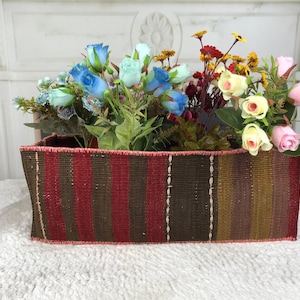 Op de afbeelding: Een rechthoekige gevlochten mand met verticale strepen in donkerbruin, dieprood en olijfgroen. De mand is gevuld met een arrangement van kunstbloemen, waaronder lichtblauwe, crèmekleurige en roze rozen naast kleine gele bloemetjes.