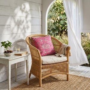 May include: A rattan armchair with a pink patterned cushion and a beige seat cushion. A small white side table holds a bowl of figs, a glass of water, and a book. The chair sits on a woven jute rug.