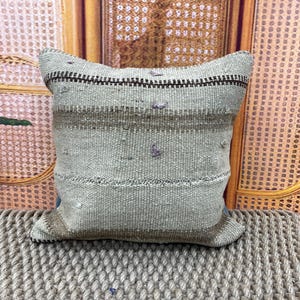 May include: A square, woven pillow with a light green and brown striped pattern. The pillow has a textured appearance with small, raised details and a dark brown border. It rests on a woven surface, suggesting a rustic or bohemian style.