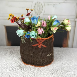 May include: A brown woven basket with a floral pattern, filled with a bouquet of artificial flowers. The flowers are a mix of light blue, pink, and yellow roses, and small yellow flowers.