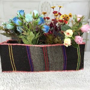 Puede incluir: Una cesta rectangular tejida con base negra y rayas verticales en amarillo, verde, rojo y azul, rematada con un borde cosido en rosa. La cesta está llena de un surtido de flores artificiales en tonos azules, rosas y amarillos.