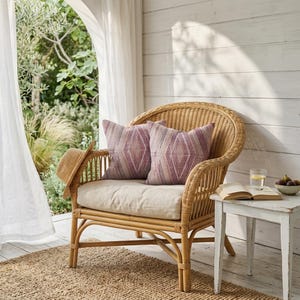 May include: A wicker armchair with two patterned throw pillows in shades of pink and white. A straw hat rests on the arm. A small white side table holds a glass of water, a bowl of figs, and an open book. The chair sits on a woven rug.