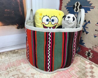 May include: A patterned storage basket with a striped design in orange, green, and brown. The basket has white handles and contains a yellow SpongeBob SquarePants plush and a panda plush. The basket is on a patterned rug.