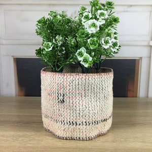 Può includere: Un vaso bianco intrecciato con un motivo geometrico, riempito di piante verdi a foglia e fiori bianchi.