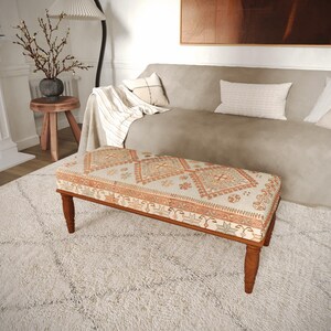 May include: A rectangular upholstered bench featuring a cream-colored fabric top with a repeating orange and brown geometric diamond pattern, supported by four turned wooden legs in a medium brown finish.