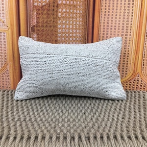 May include: A rectangular decorative pillow with a textured, light gray and black woven design. The pillow is placed on a woven surface. The background features a light brown, woven pattern.