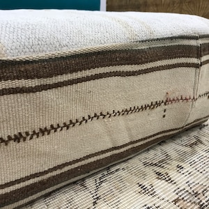 May include: A white and brown striped floor cushion with a stitched design. The cushion is made of woven fabric and has a rectangular shape.