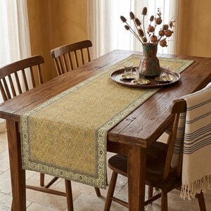 a wooden dining table with a yellow and green patterned runner, a tray with glasses, and a vase of dried flowers on it. The table is surrounded by wooden chairs, and there is a window in the background.