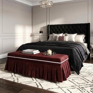 May include: A luxurious bedroom scene featuring a black tufted headboard, a bed with black bedding, and a burgundy ottoman with a pleated skirt. A gold-framed mirror and a patterned rug add elegance. A candle and books are on the ottoman.