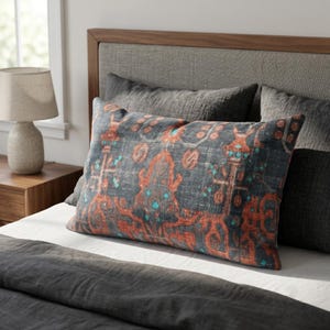 May include: A rectangular throw pillow featuring a dark charcoal gray base with a prominent, intricate pattern in burnt orange and bright turquoise. The design displays symmetrical, ornate motifs across the fabric surface, centered on a bed.