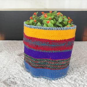 May include: A cylindrical woven fabric basket featuring horizontal stripes in vibrant yellow, red, purple, and blue. The basket is filled with a small artificial plant that has green leaves and tiny orange flowers, creating a colorful display.