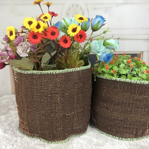 May include: Two brown fabric plant pots with green trim. The pot on the left has a bouquet of yellow, red, and blue flowers. The pot on the right has a green plant with small red flowers.