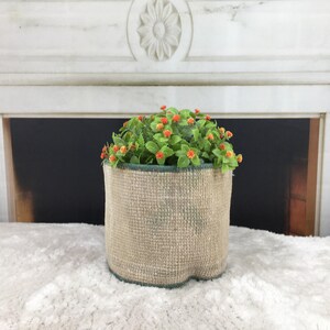 May include: A cylindrical planter with a textured, light tan woven fabric exterior and a dark green rim. The planter contains a dense cluster of artificial green foliage featuring small, round, bright orange flower buds throughout the plant.