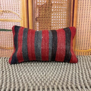 May include: A rectangular throw cushion featuring a woven textile pattern with vertical stripes in shades of deep red, charcoal grey, and black. The fabric has a coarse, textured appearance with visible thread variations throughout the surface.