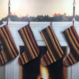 May include: Four striped Christmas stockings hang on a white mantel above a fireplace. The stockings are brown, orange, and red with white trim.