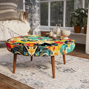 Op de afbeelding: Een ronde poef met een levendig, veelkleurig bloemen- en geometrisch patroon in tinten geel, taling, rood en zwart. Het meubelstuk heeft een gestoffeerde bovenkant en vier taps toelopende houten poten voor een lage zit- of tafeloptie.