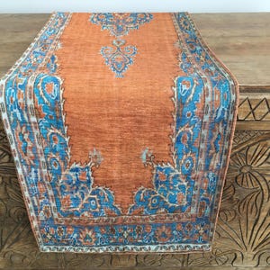 May include: A long orange table runner with a blue and white patterned border. The runner has a traditional design with intricate floral motifs.