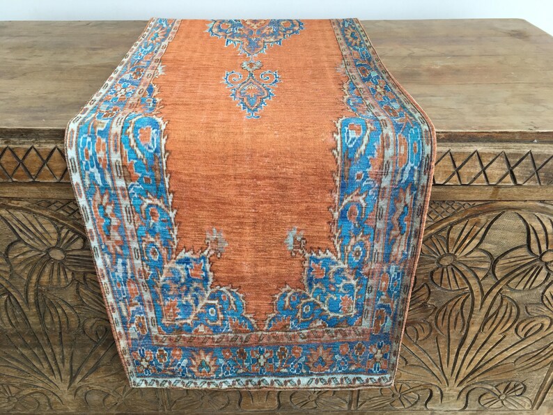 May include: A long orange table runner with a blue and white patterned border. The runner has a traditional design with intricate floral motifs.