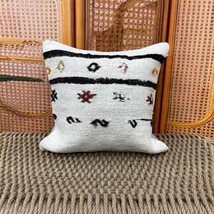 May include: A white decorative pillow with a woven texture. The pillow features black, brown, and red geometric patterns. The pillow is placed on a woven surface, with a woven background.