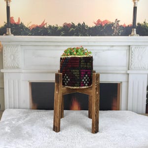 Peut inclure: Un cache-pot cylindrique en tissu tissé avec un motif géométrique multicolore foncé, contenant une petite plante verte, posé sur un support en bois à quatre pieds avec une finition teintée foncée et un grain de bois visible.