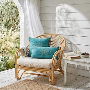 Puede incluir: Una silla de ratán natural con un cojín color crema y dos cojines decorativos turquesa con un patrón oscuro. Una pequeña mesa auxiliar blanca tiene un libro, un vaso de agua y un cuenco con higos. Una manta de color neutro está sobre el brazo.