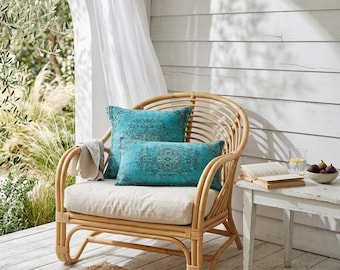 May include: A natural rattan chair with a cream-colored cushion and two turquoise decorative pillows with a dark pattern. A small white side table holds a book, a glass of water, and a bowl of figs. A neutral-colored throw is draped over the arm.