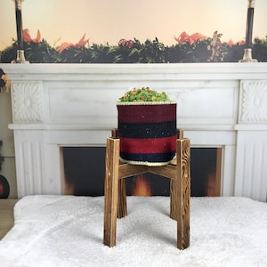 Pode incluir: Um suporte de plantas de madeira com quatro pernas, que suporta um vaso tecido com uma planta verde e bagas vermelhas. O suporte está na frente de uma lareira branca com uma guirlanda verde.