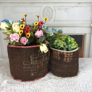 Puede incluir: Dos cestas cilíndricas de tejido marrón oscuro con superficies texturizadas. La cesta más grande presenta un borde cosido de color coral y contiene un ramo de flores artificiales rosas, amarillas, azules y rojas. La pequeña tiene un borde crema.